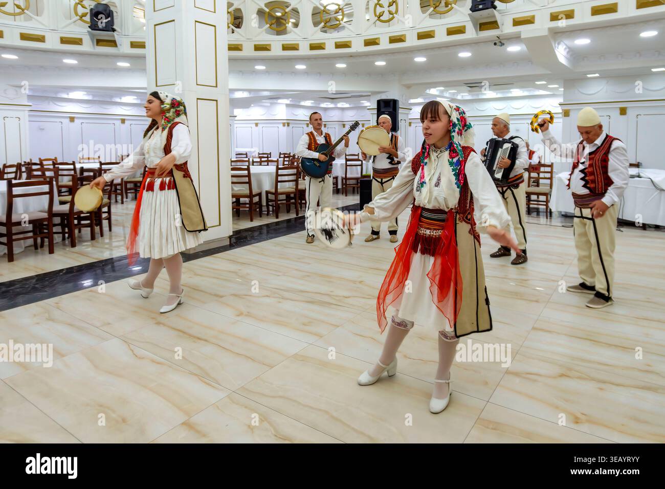 Gruppo tradizionale albanese che esegue danza e musica in una sala banchetti con artisti in costumi etnici e strumenti Foto Stock