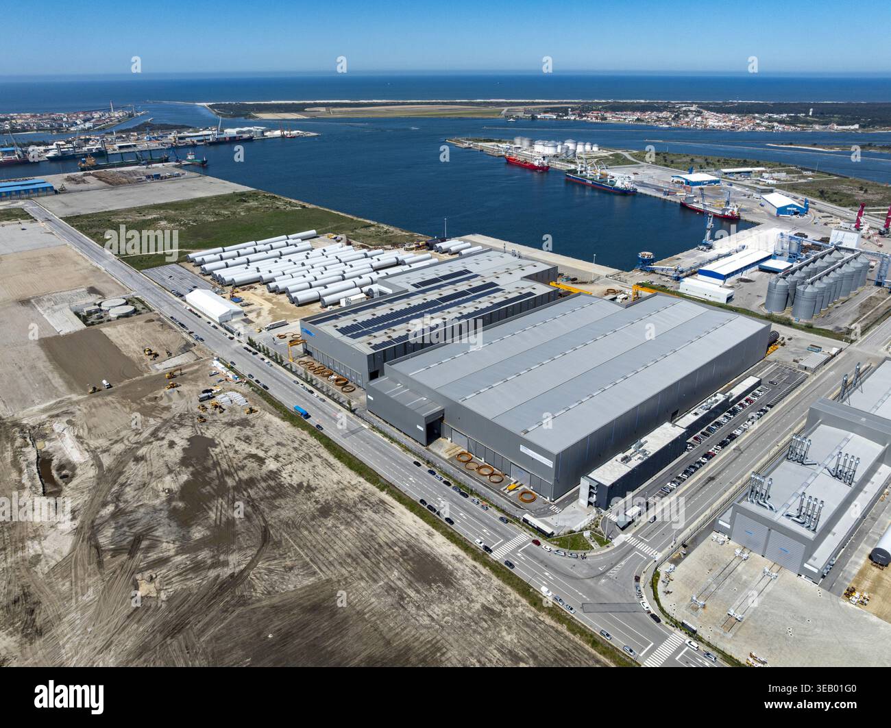 Vista aerea dell'ampio complesso industriale con i suoi tetti metallici che riflettono il cielo luminoso, contrastato dal blu profondo delle acque del porto, A. Foto Stock