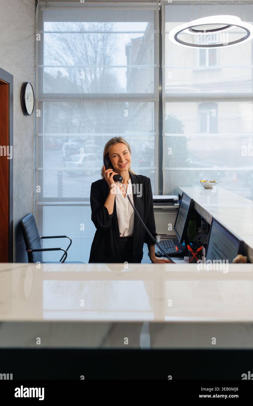 La receptionist dell'hotel è cordiale e parla al telefono dietro la reception nella hall moderna Foto Stock