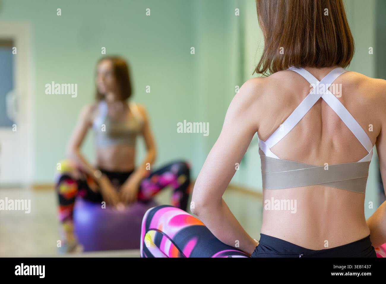 Vista posteriore di una donna in forma con capelli corti che indossa un reggiseno sportivo e leggings colorati in palestra, con una lezione di esercizi sfocati sullo sfondo. Messa a fuoco o Foto Stock
