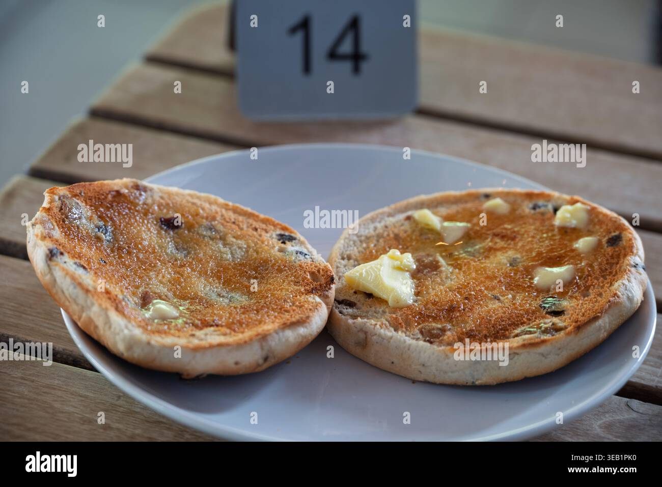 Le metà del panino di frutta tostato dorato, brillanti con burro fuso, sono presentate su un piatto bianco al tavolo 14. Foto Stock
