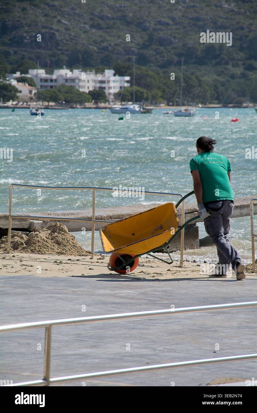 Mallorca Spagna - 24 aprile 2026: Un operaio manuale in una polo verde di servizio tecnico spinge una carriola gialla lungo un sentiero costiero a Port de po Foto Stock