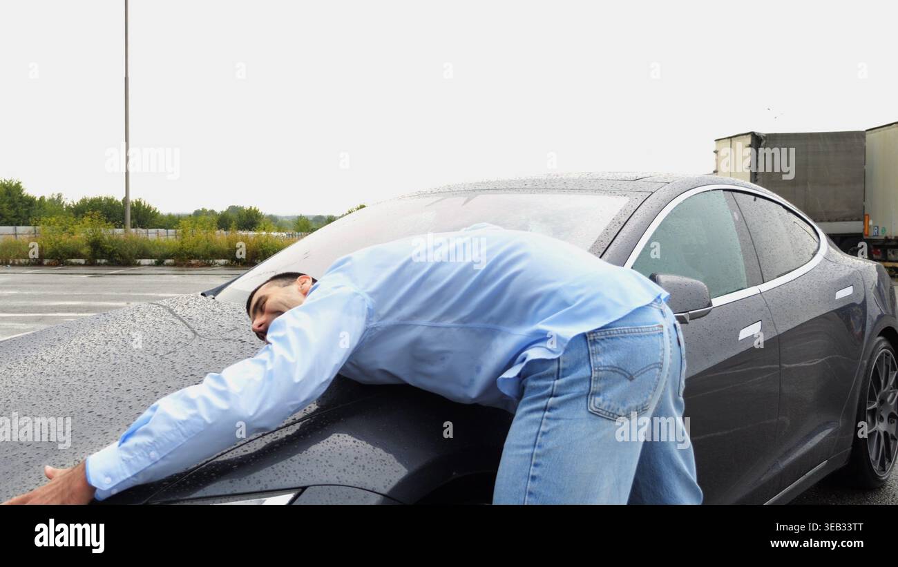 Un imprenditore sorridente di successo acquistò un nuovo veicolo elettrico e avvolge il cofano dell'automobile. Giovane uomo d'affari felice in camicia che ama il moderno Foto Stock
