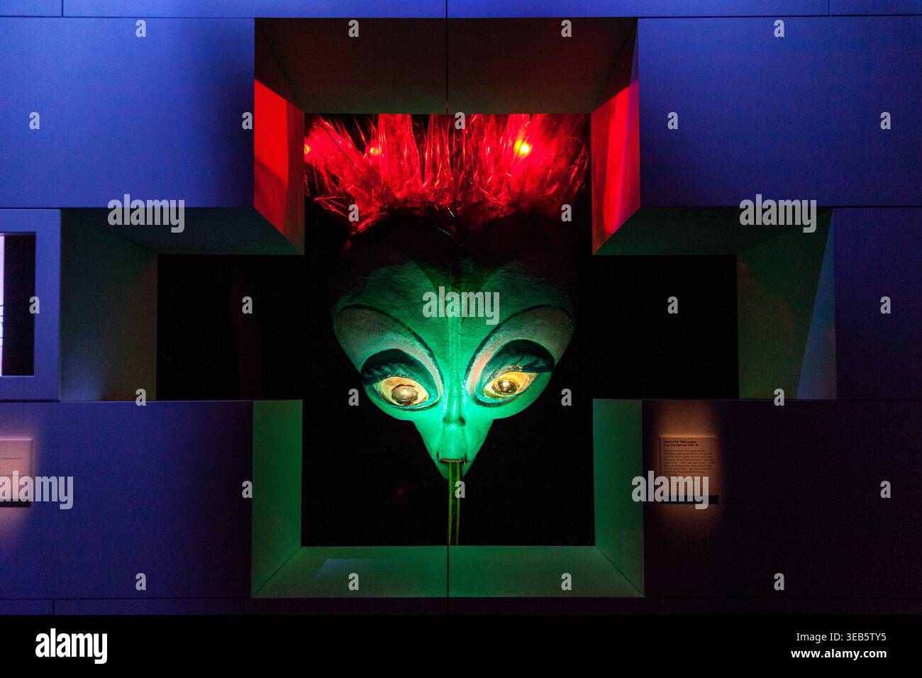 Scultura verde illuminata con occhi gialli brillanti, incorniciata da luci al neon rosse su sfondo architettonico blu scuro, mostra Pink Floyd Foto Stock