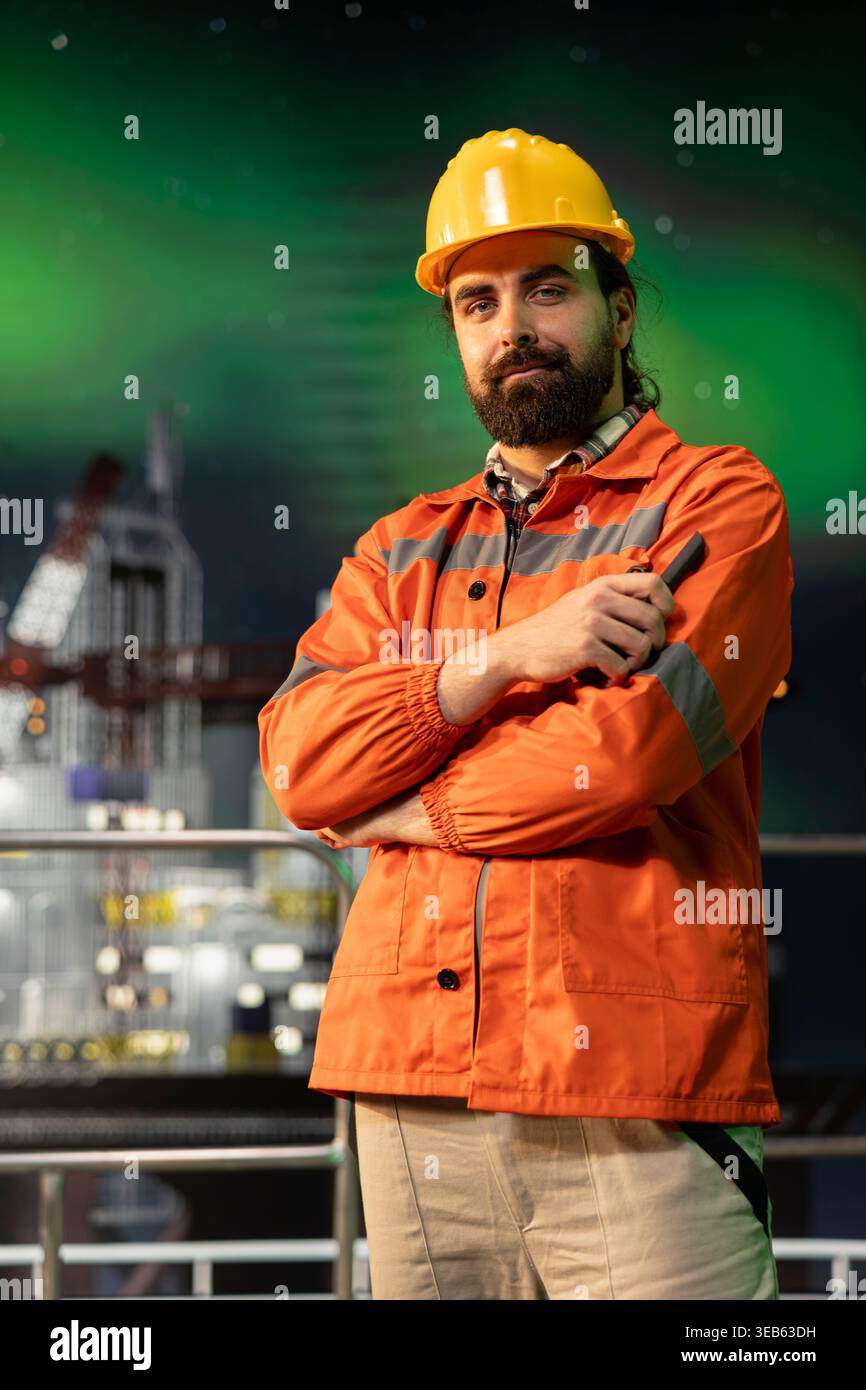 Ritratto di un tecnico della piattaforma offshore sorridente che supervisiona le operazioni durante la notte dell'aurora boreale. Lo specialista di carri di perforazione Happy supervisiona le attività di produzione sul ponte, sfondo verde dell'aurora boreale Foto Stock