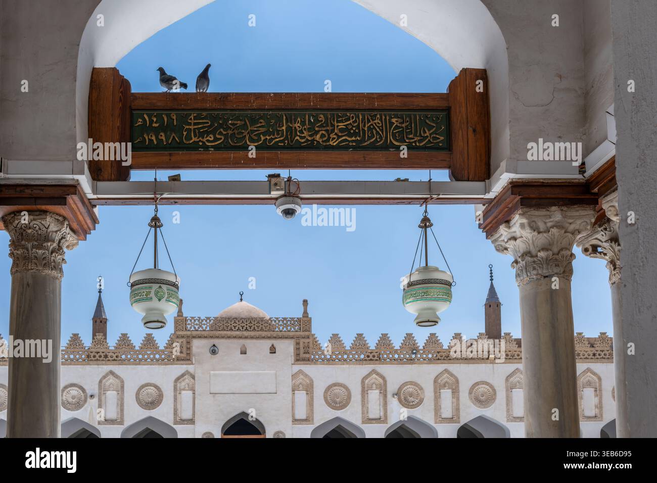 Vista della storica Moschea al-Azhar con i suoi archi bianchi ornati, lanterne appese e piccioni arroccati su una trave di legno sotto un cielo azzurro e limpido il Cairo Foto Stock