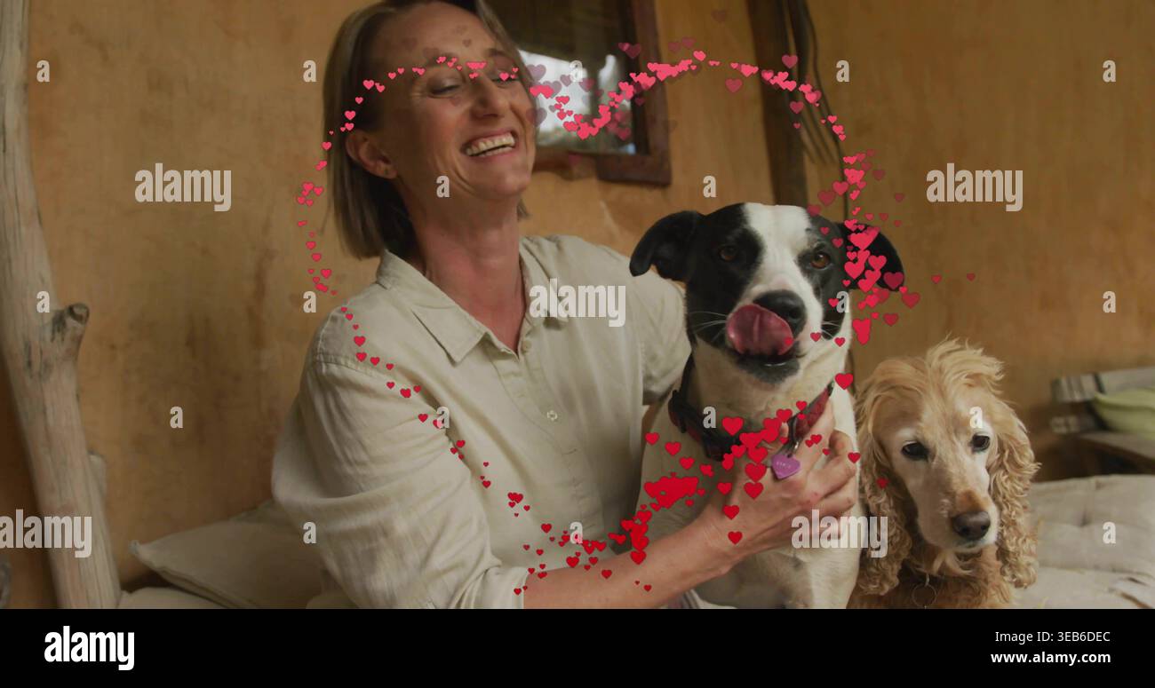 Donna sorridente in camicia beige che tiene due cani sul divano nel soggiorno, copricuore e collari Foto Stock
