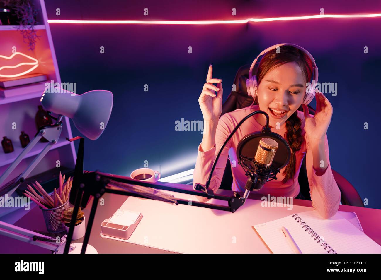 Canale ospite della giovane bella emittente asiatica che ascolta la musica sui social media con gli ascoltatori, indossa cuffie pastello e usa il microfono per Foto Stock