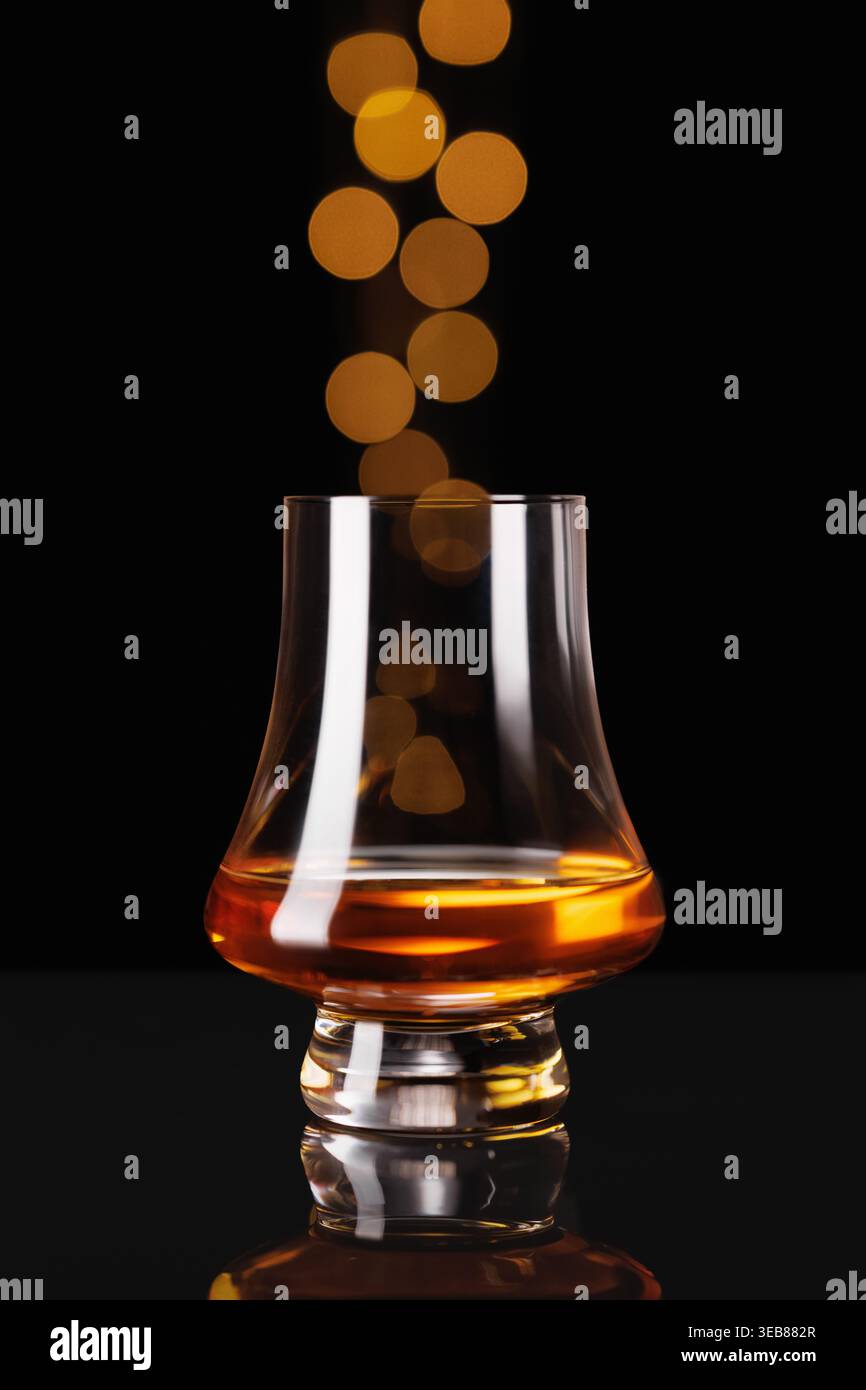 Bicchiere di whisky riempito di liquido ambrato che riflette calde luci bokeh dorate su sfondo scuro Foto Stock