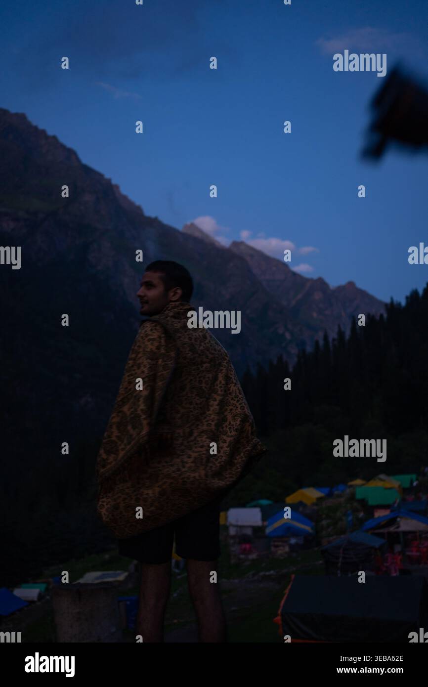 Si erge un uomo solitario, ammirando il tranquillo paesaggio montano e il lontano e colorato villaggio annidato nella valle mentre il crepuscolo si deposita sul maestoso Foto Stock