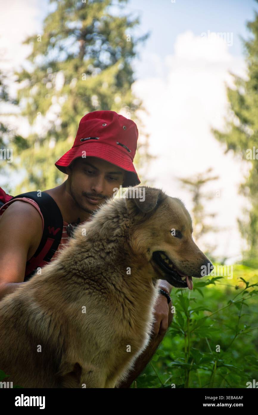 Questo primo piano cattura un uomo allegro con un cappello rosso che guarda con affetto il suo cane contento durante una rinfrescante escursione all'aperto in una foresta soleggiata a Jibh Foto Stock