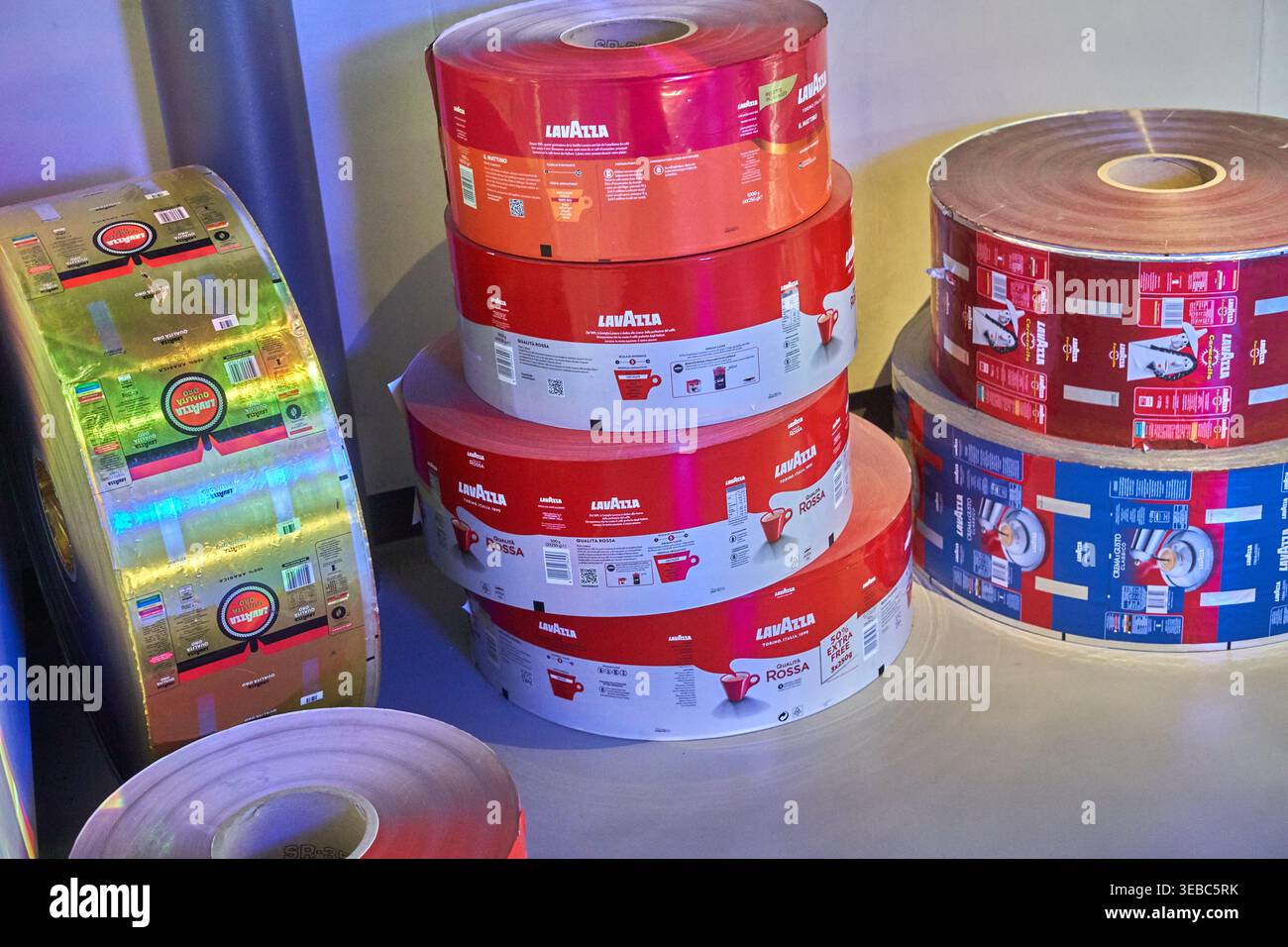 Grandi rotoli cilindrici di film per imballaggio flessibile stampato impilati in uno stabilimento di produzione industriale. La stampa rossa e bianca prominente Foto Stock