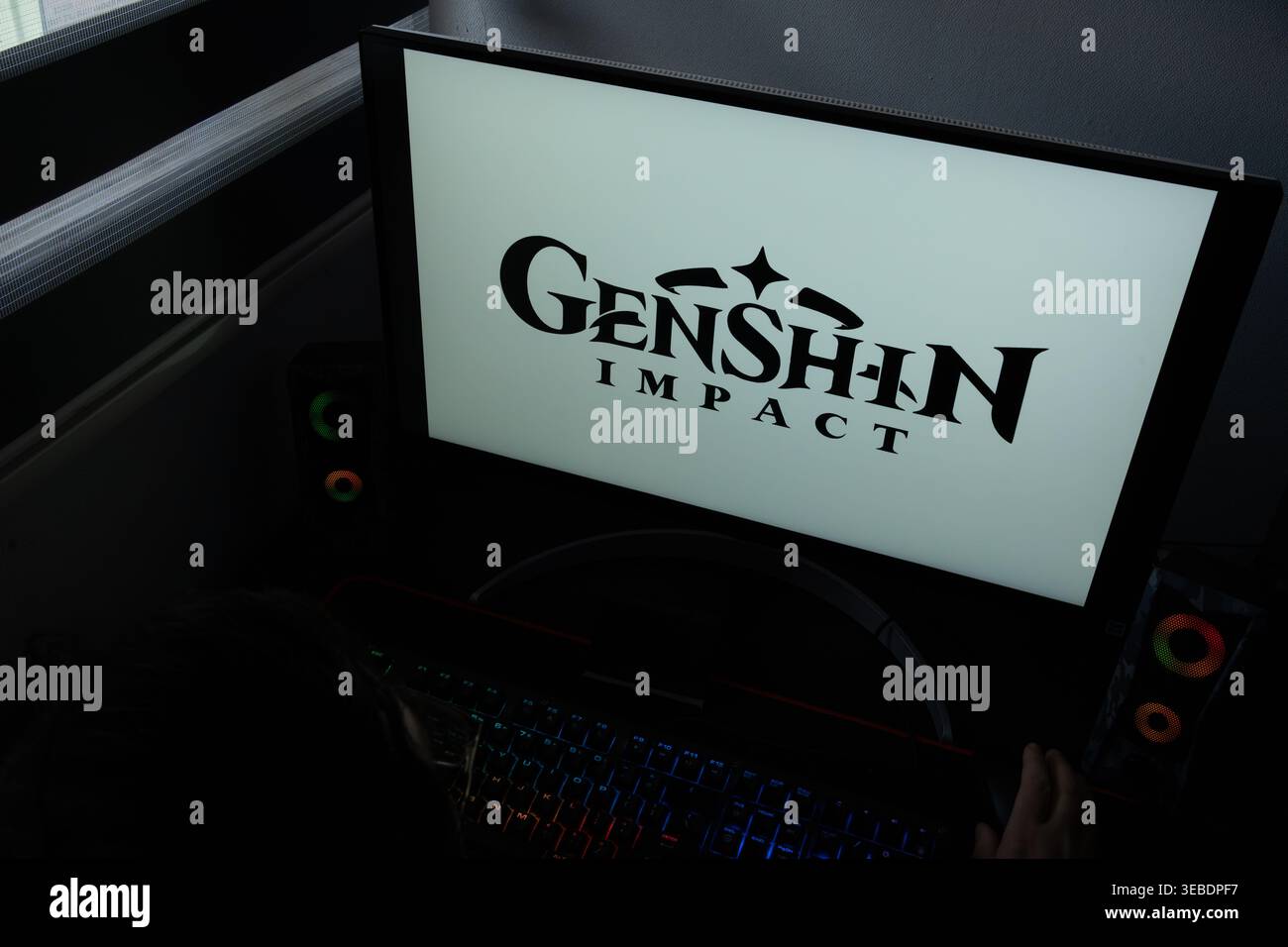 Il logo del videogioco Genshin Impact viene visualizzato sul monitor di un computer in un ambiente di home Office scarsamente illuminato Foto Stock