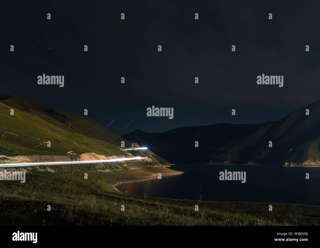 Le piste luminose dei veicoli illuminano una tortuosa strada di montagna accanto a un lago sereno sotto un cielo notturno buio Foto Stock