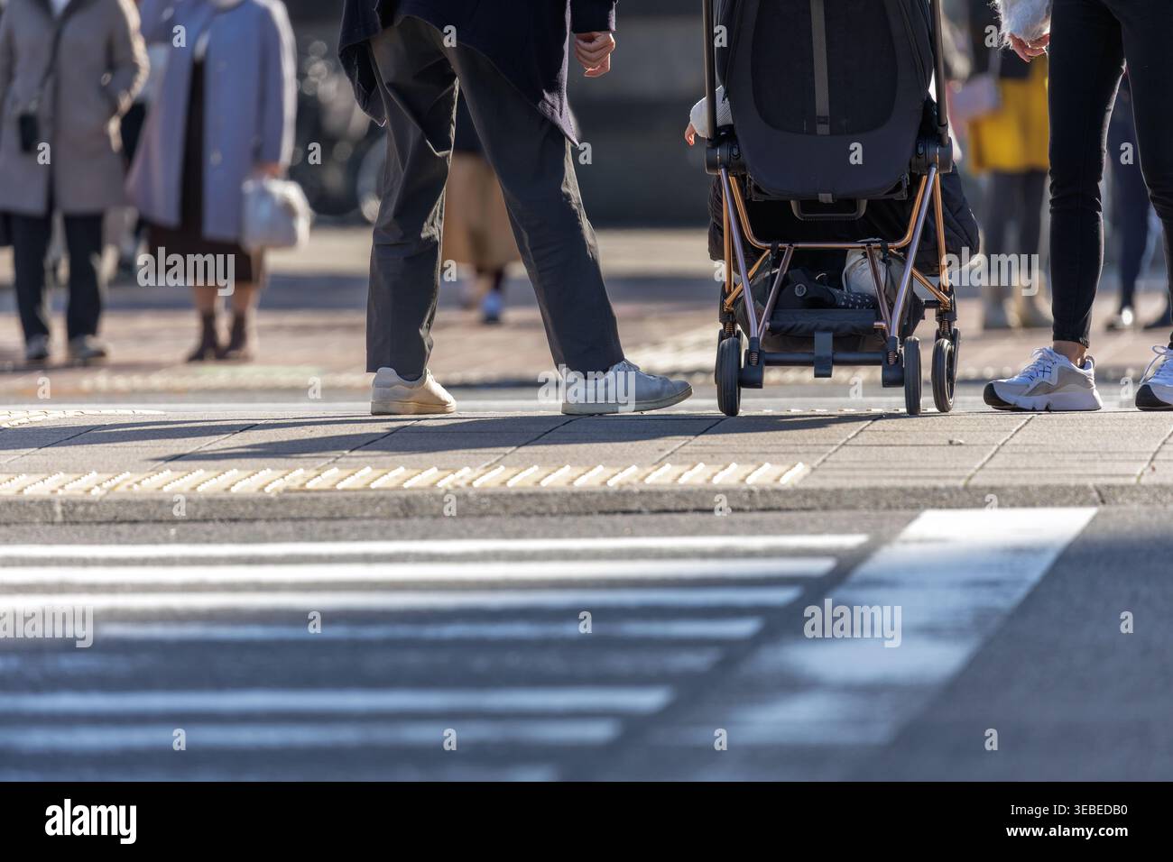 Angolo inferiore di coppia con bambino nel passeggino in attesa sulla mediana centrale con pedoni sullo sfondo bokeh in attesa di attraversare dal lato opposto. Foto Stock