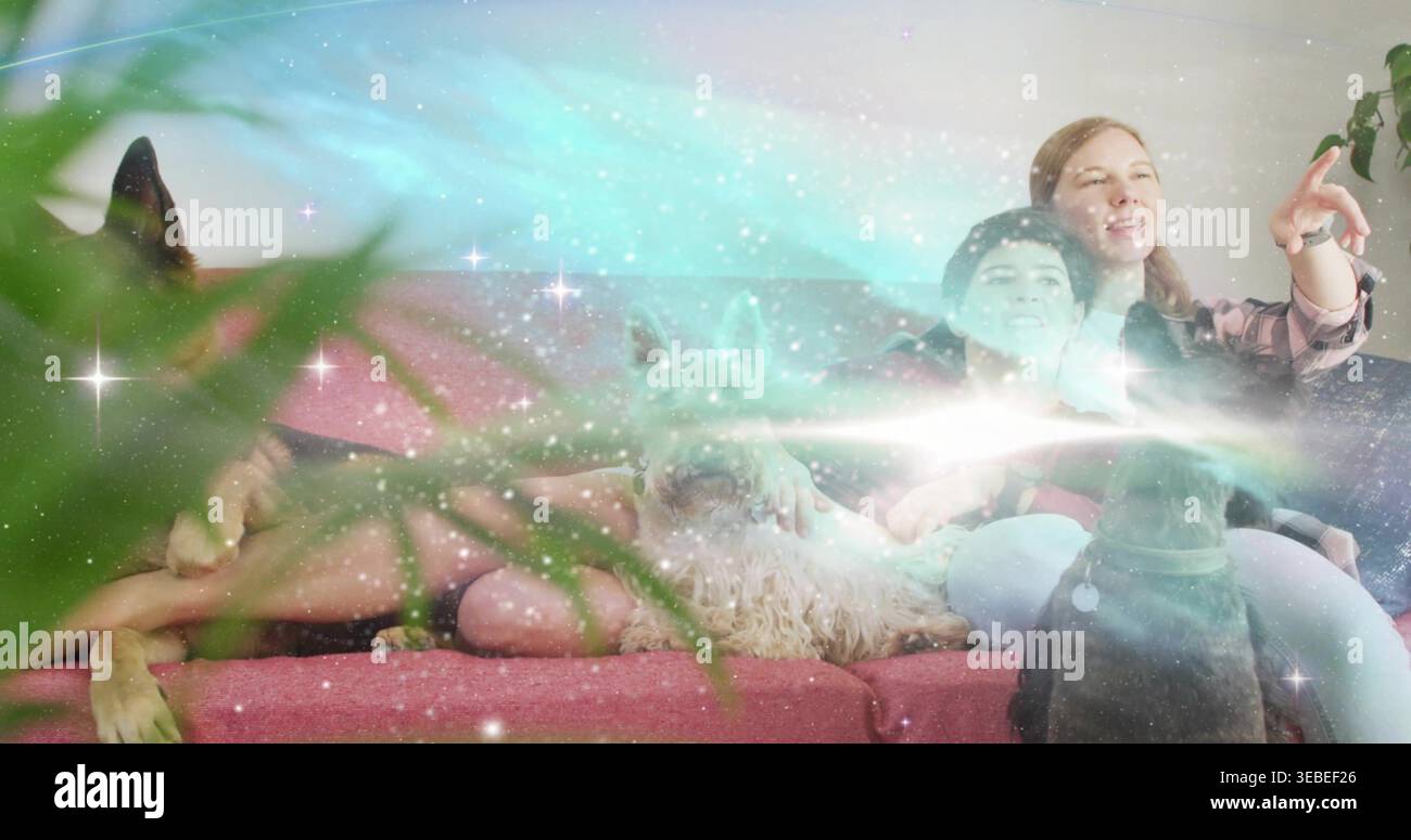 Donne sorridenti sedute sul divano rosa a casa, indossano camicia a quadri, cani da compagnia, piante e aurora Foto Stock