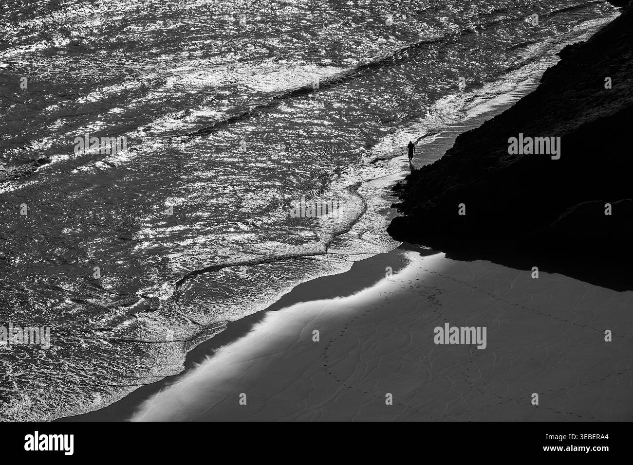 Solitario che cammina su una spiaggia remota in Portogallo, con onde, scogliere e scintillanti coste atlantiche in bianco e nero. Foto Stock