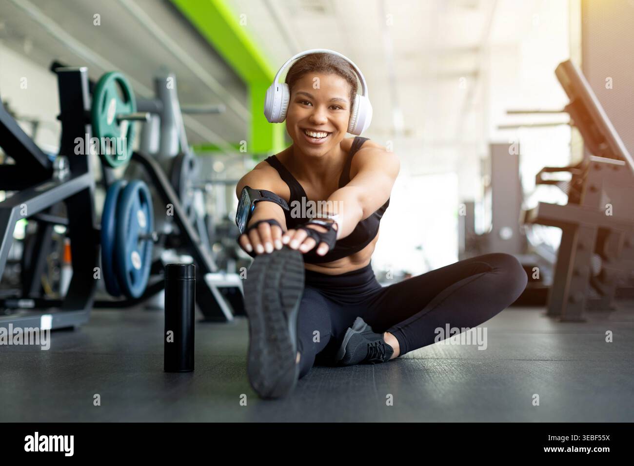 Sorridente giovane donna nera seduta sul pavimento della palestra allungando le gambe in avanti Foto Stock