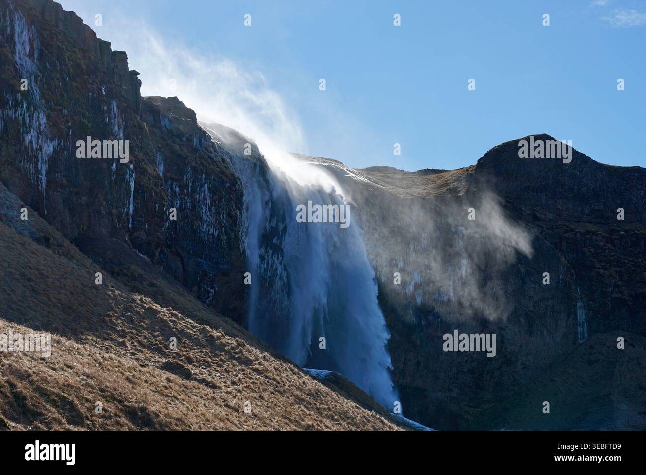 Potente cascata di scogliera con spray nebulizzato e prugne soffiate dal vento nel paesaggio montano illuminato dal sole Foto Stock