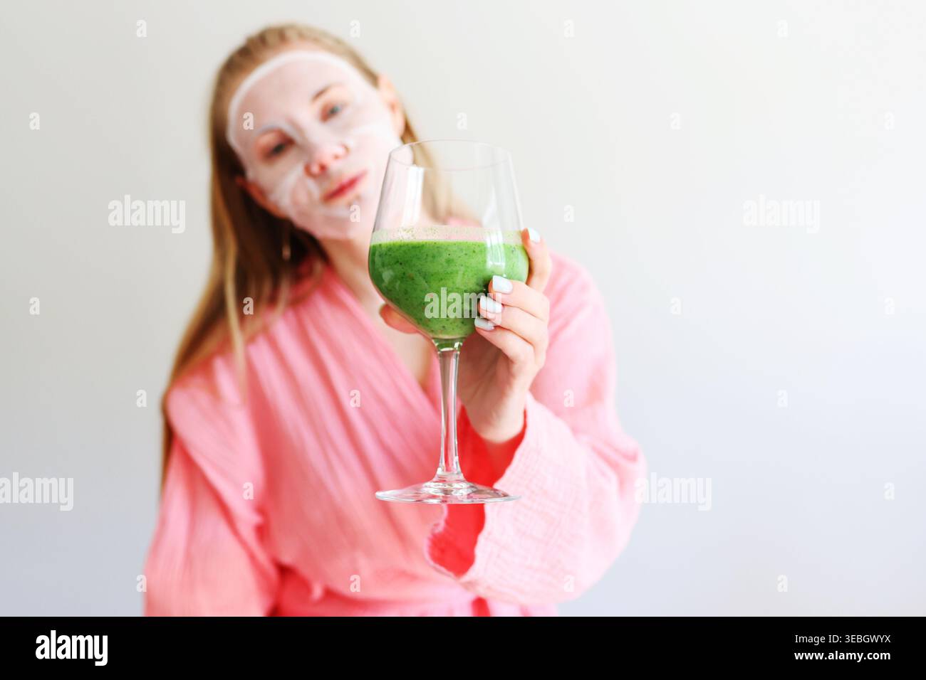 Centro benessere di bellezza a casa, donna in accappatoio con maschera viso e succo verde, concetto di cura della pelle. Foto Stock