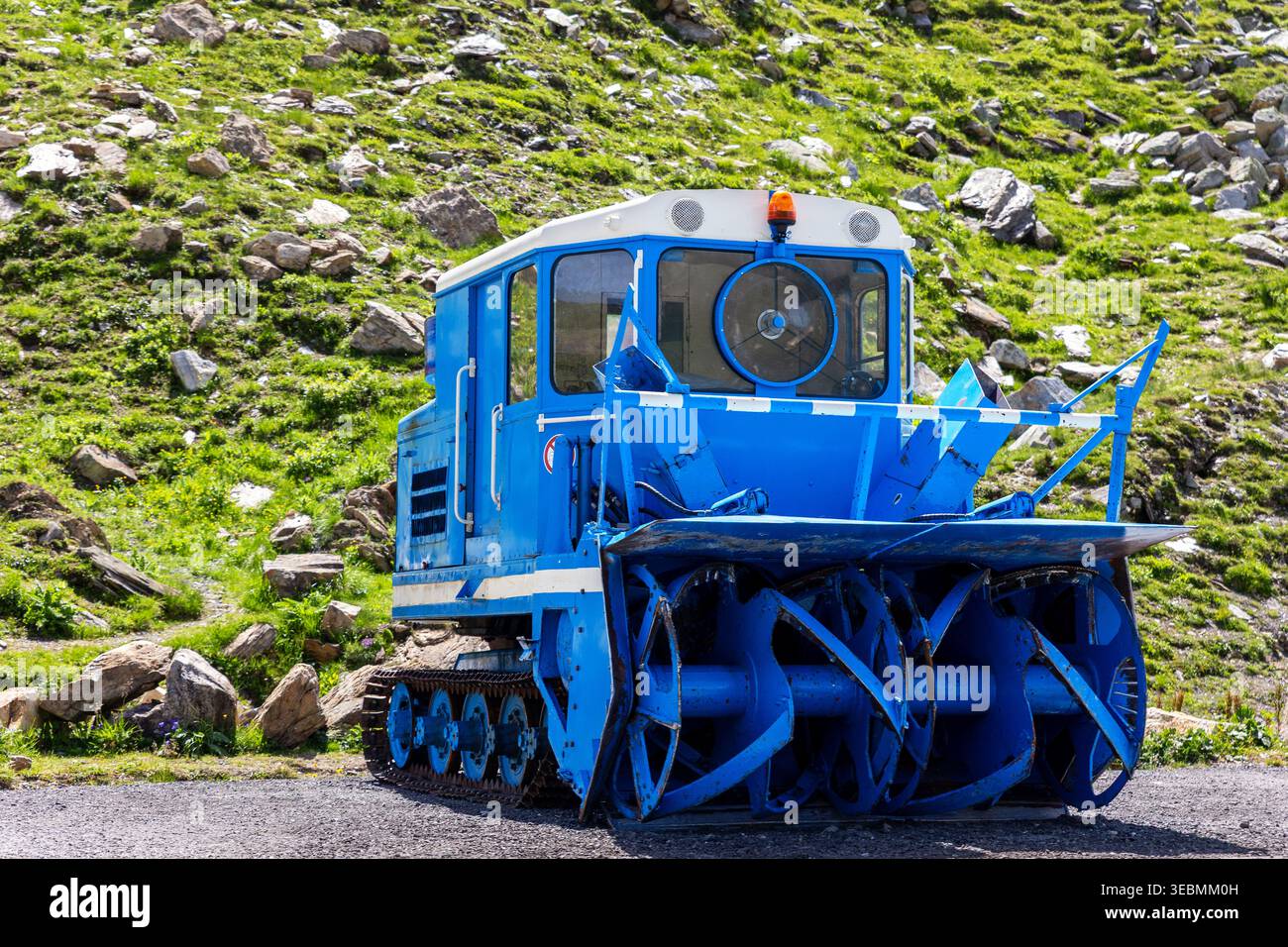 Macchina per spazzaneve blu grandi lame rotanti si staglia su strada ghiaia paesaggio alpino rocce di erba verde. Rimozione della neve progettata per i veicoli industriali cingolati Foto Stock