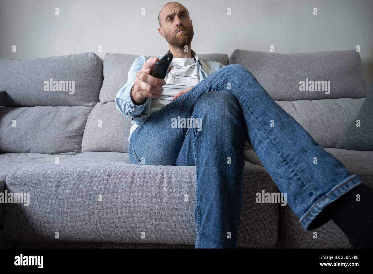 Uomo seduto comodamente su un divano di casa, con un telecomando e guardando la televisione. Ambiente interno naturale con luce soffusa che rappresenta il leisur Foto Stock