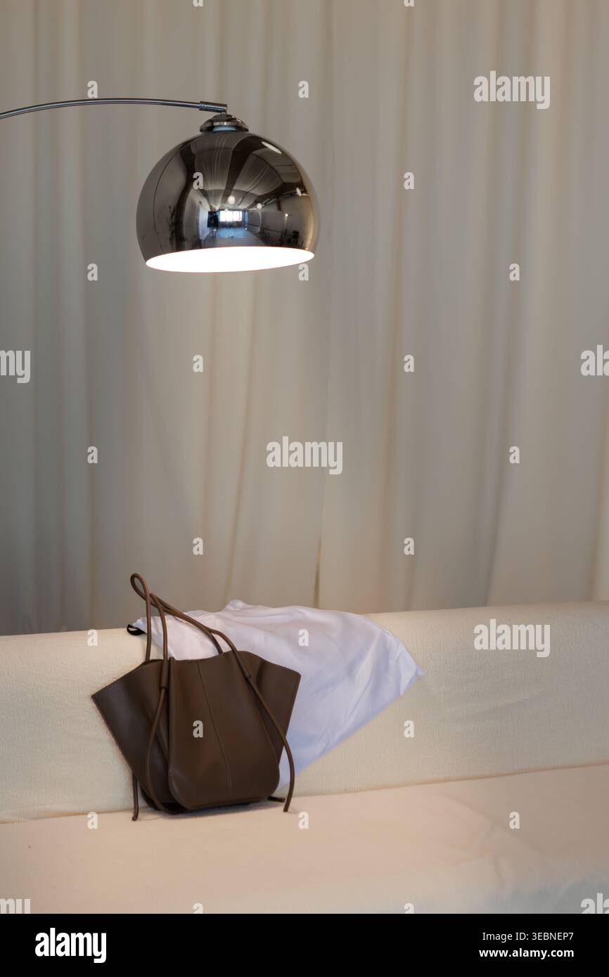 Una lampada emette luce sopra un divano dove una borsa marrone si trova accanto a un pezzo di tessuto bianco. La scena mostra un intimo spazio interno con colori tenui e m Foto Stock