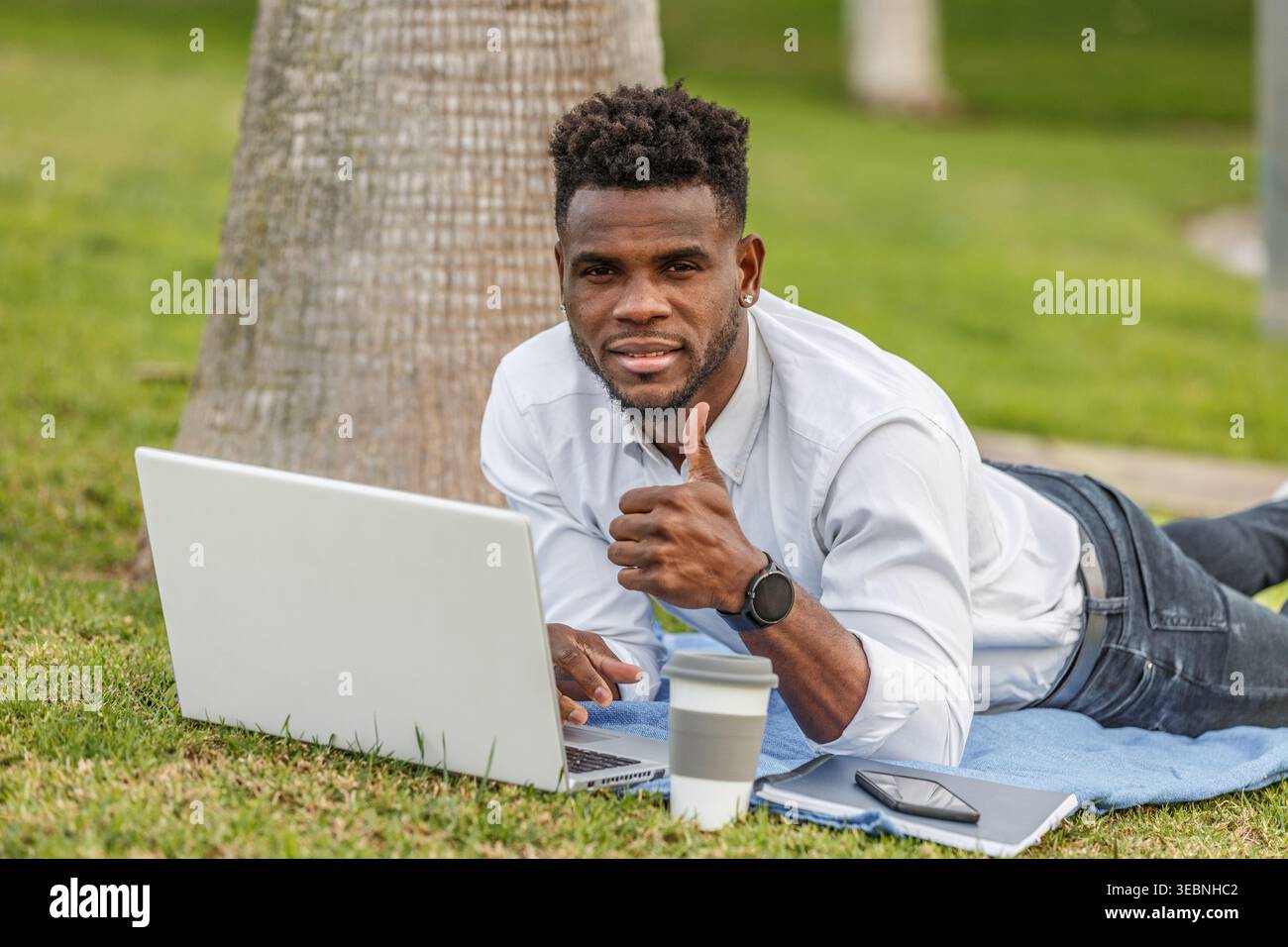 Un uomo afroamericano viene visto con il pollice sdraiato sul pavimento mentre utilizza un computer portatile per lavorare. Foto Stock