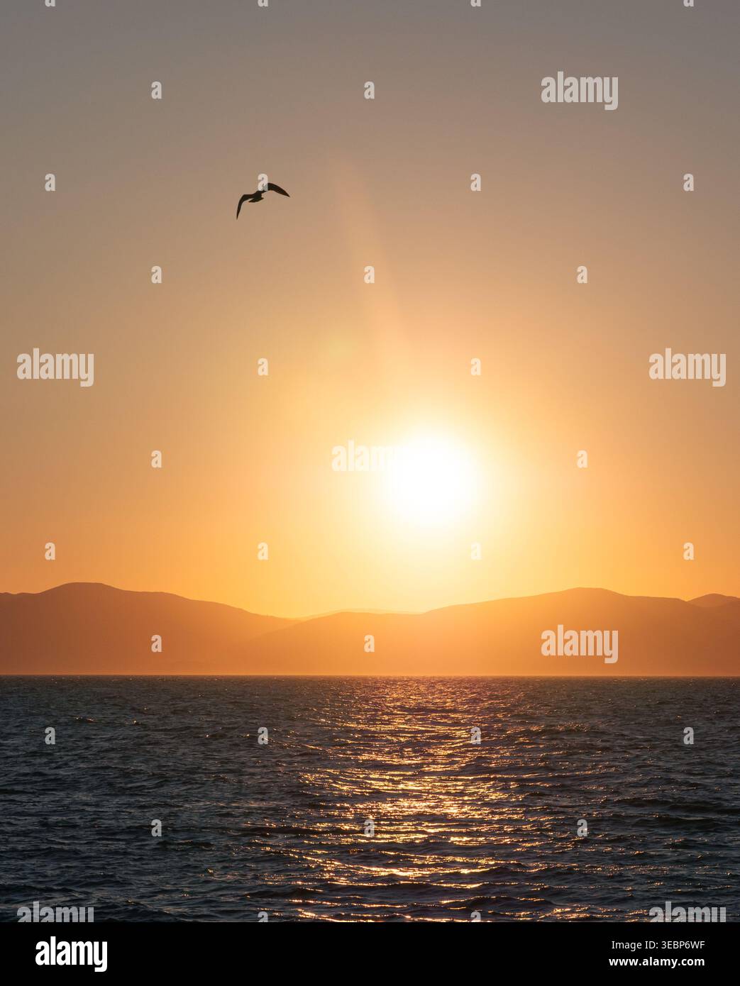 Il sole si tuffa sotto montagne lontane dipingendo il vasto oceano con un caldo e ardente bagliore al crepuscolo Foto Stock