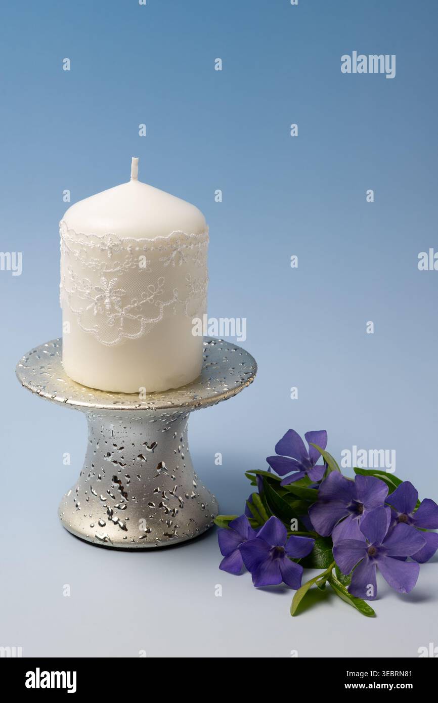 Candela decorativa bianca su supporto in metallo e fiori viola su sfondo blu con spazio di copia Foto Stock