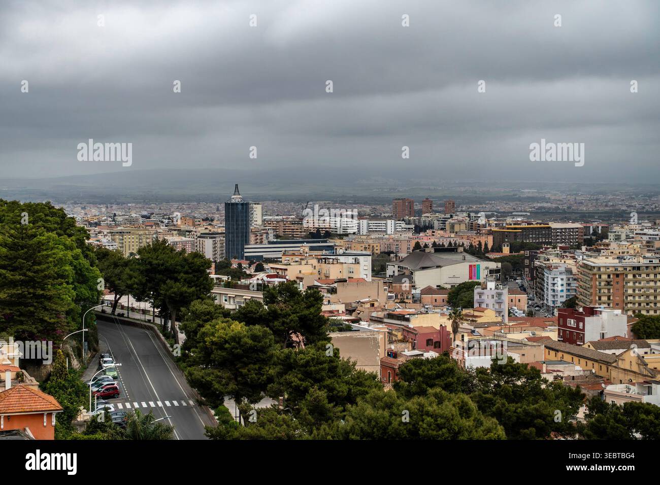 Una veduta sopraelevata della proliferazione urbana di Cagliari sotto un pesante cielo coperto. Le cime degli alberi e una strada tortuosa incorniciano la città e le colline lontane. Foto Stock