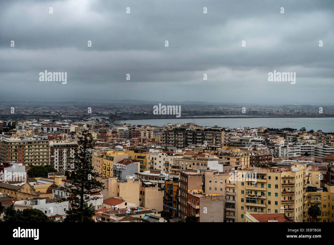 Una vista ad alto angolo di Cagliari che mostra dense abitazioni urbane e la laguna dello stagno di Cagliari sotto un cielo pesante e coperto di nuvole scure. Foto Stock