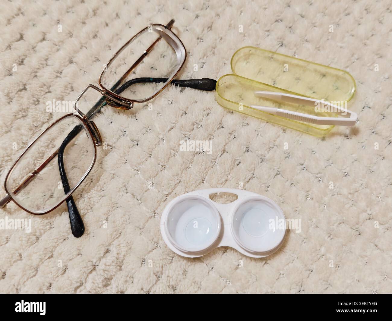 Occhiali, una custodia per lenti a contatto e pinzette poggiano su un tappeto di colore chiaro in un ambiente domestico. Questi elementi mostrano la routine quotidiana di cura della vista. Foto Stock
