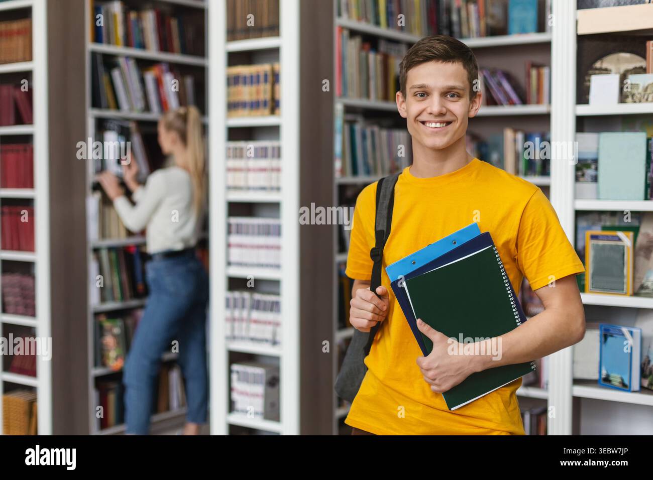 Il giovane studente sorride mentre tiene i libri in una biblioteca durante le ore di studio pomeridiane Foto Stock