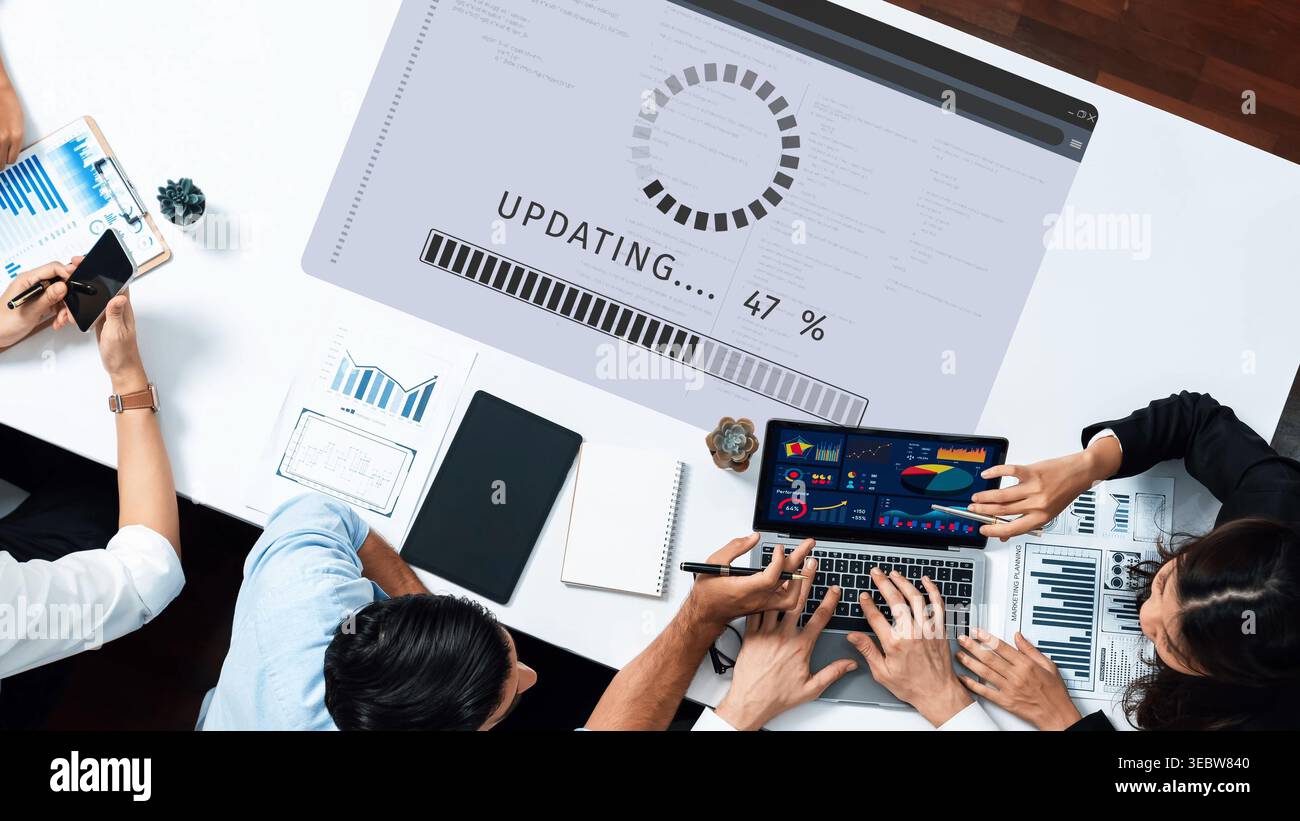 Un'area di lavoro collaborativa che mostra lo schermo di un notebook che mostra i progressi degli aggiornamenti e i grafici colorati. Individui diversi si concentrano sul digitale Foto Stock