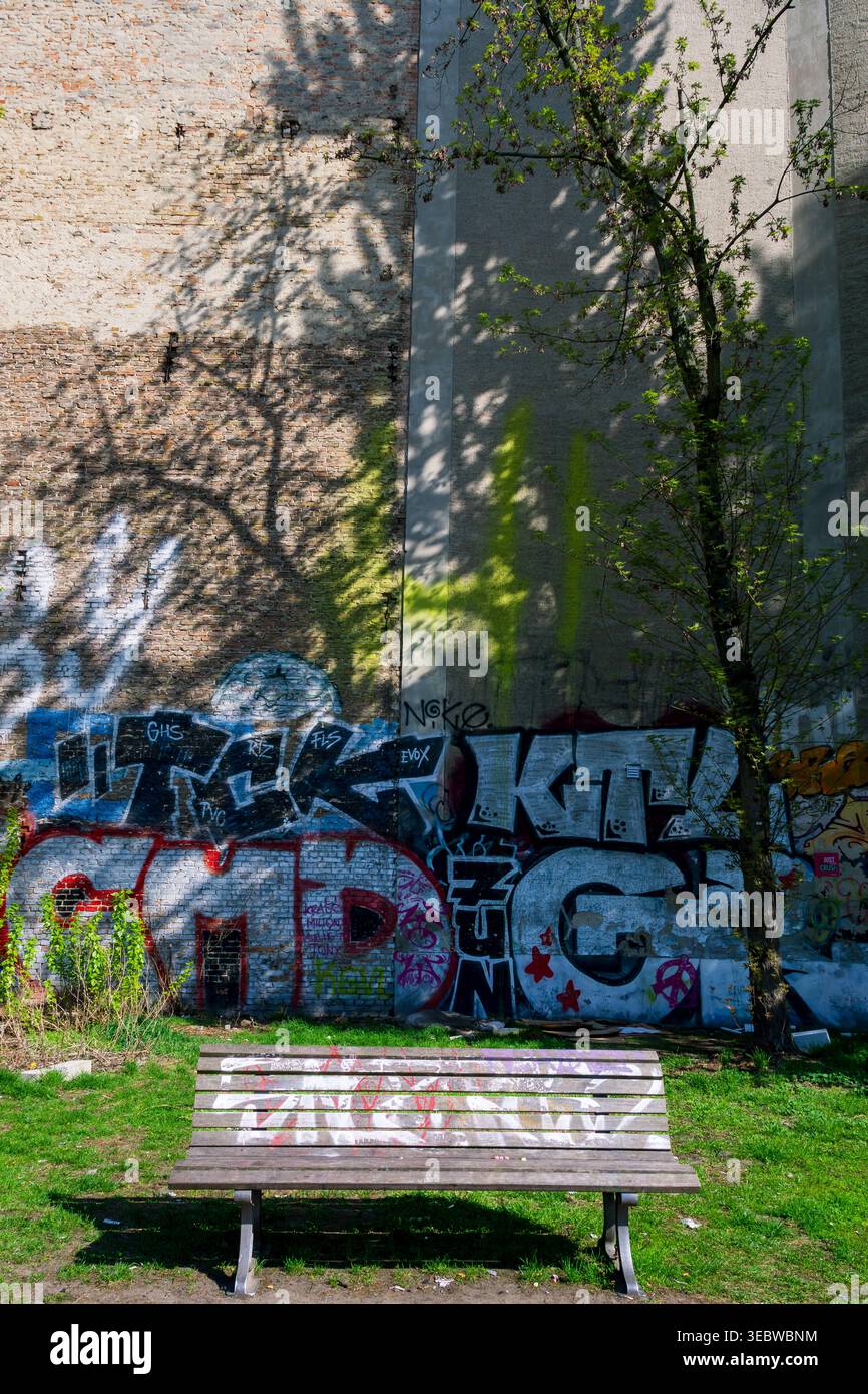 Panchina vuota di fronte a un muro coperto di graffiti nel quartiere Kreuzberg di Berlino, Germania. Foto Stock