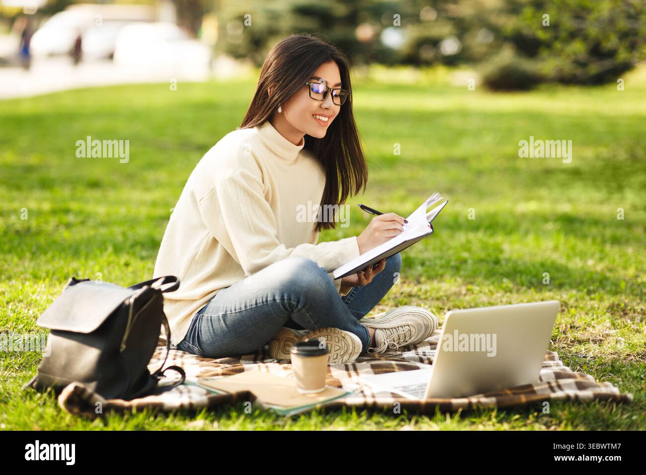 Giovane donna seduta su erba che scrive su un notebook mentre utilizza un notebook all'aperto Foto Stock