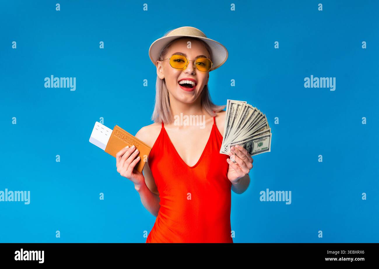 Happy Young Woman in costume da bagno in possesso di contanti e biglietto su sfondo blu Foto Stock