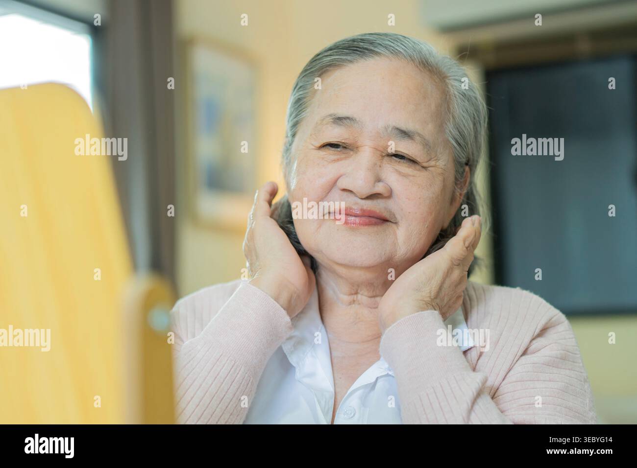 Bella donna asiatica anziana sorridente allo specchio mentre tocca la faccia. Donna sicura di sé che ammira la pelle liscia a casa. Messa a fuoco selettiva. Foto Stock