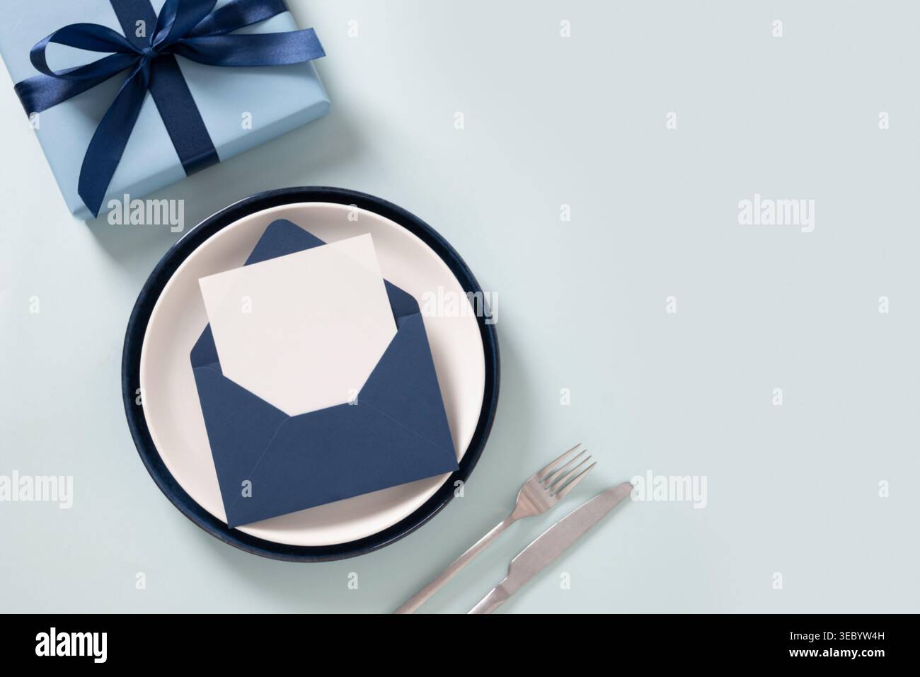 Confezione regalo blu con busta a nastro e carta bianca su piatto con posate, composizione minima su sfondo chiaro, concetto di celebrazione, spazio per copie. Grasso Foto Stock
