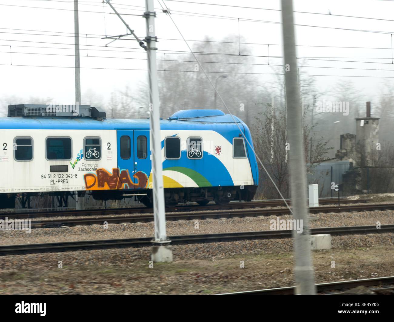 Cracovia, Polonia - 3 dicembre 2025: Un treno passeggeri blu e bianco con graffiti e simboli delle biciclette si sposta lungo i binari ferroviari in un'area industriale nebbiosa Foto Stock