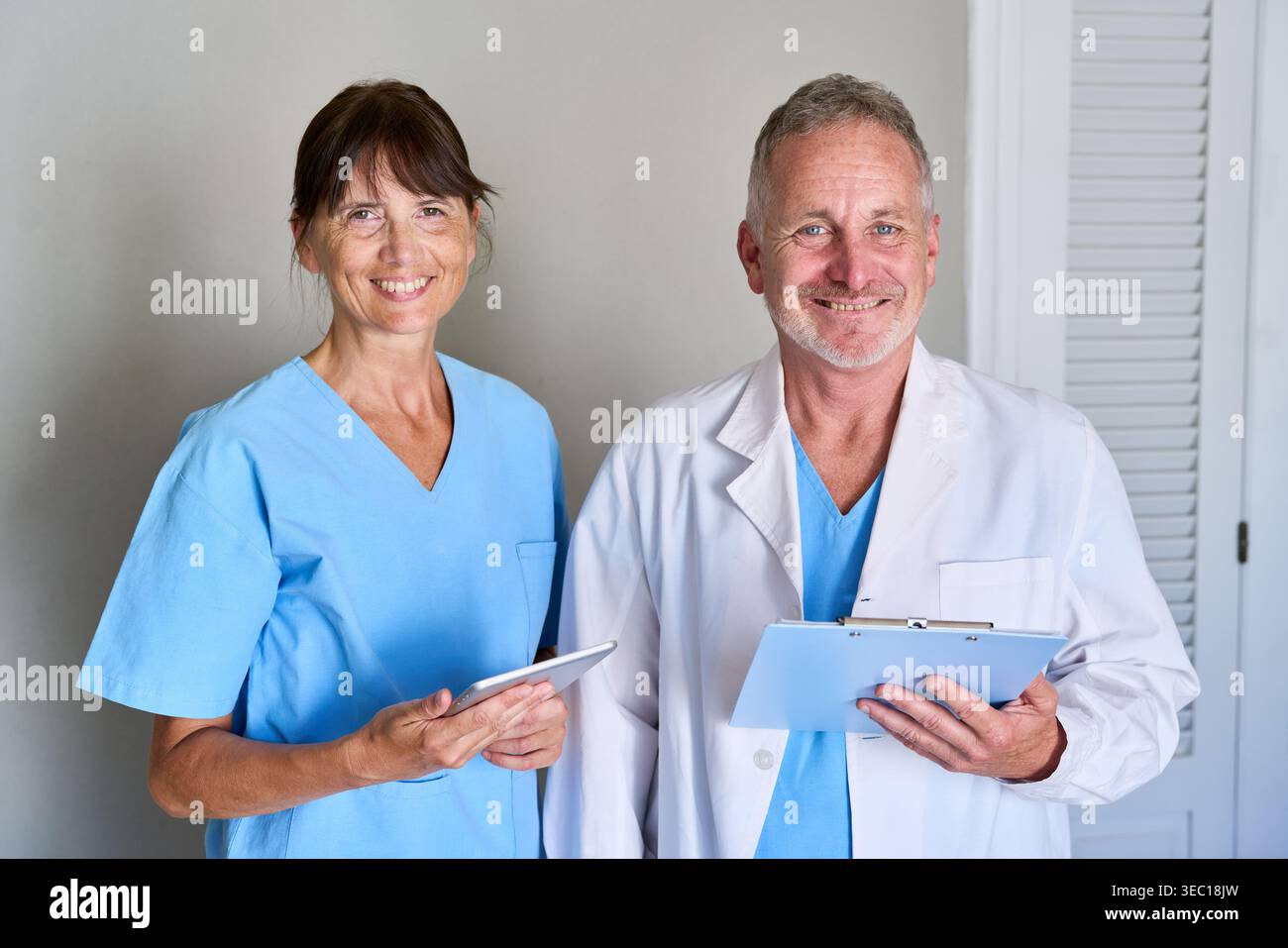 Operatori sanitari — un medico di fiducia maschile in un camice da laboratorio con un portadocumenti e un'infermiera di sesso femminile in scrub blu che tiene una compressa, entrambi sorridenti a. Foto Stock
