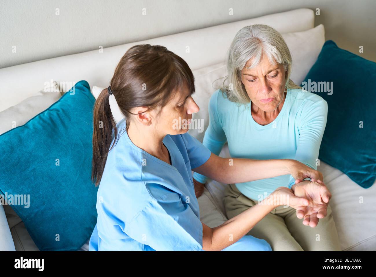 Assistenza sanitaria di alto livello — un infermiere in scrub blu sta esaminando il polso di una donna anziana, che sembra essere in disagio, durante una visita a casa. Foto Stock