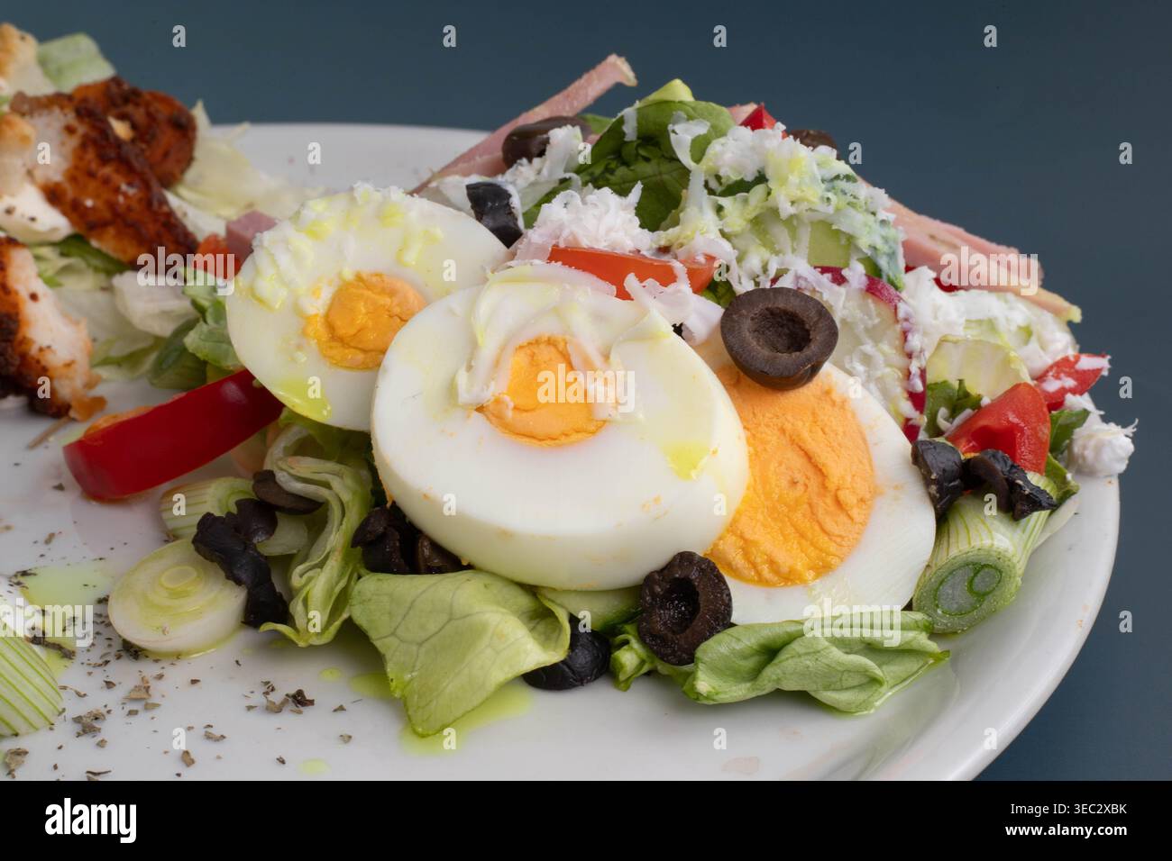 Primo piano di un'insalata fresca dello chef con fette di uova sode, olive nere, cipolle verdi a fette e prosciutto su un piatto bianco. Foto Stock