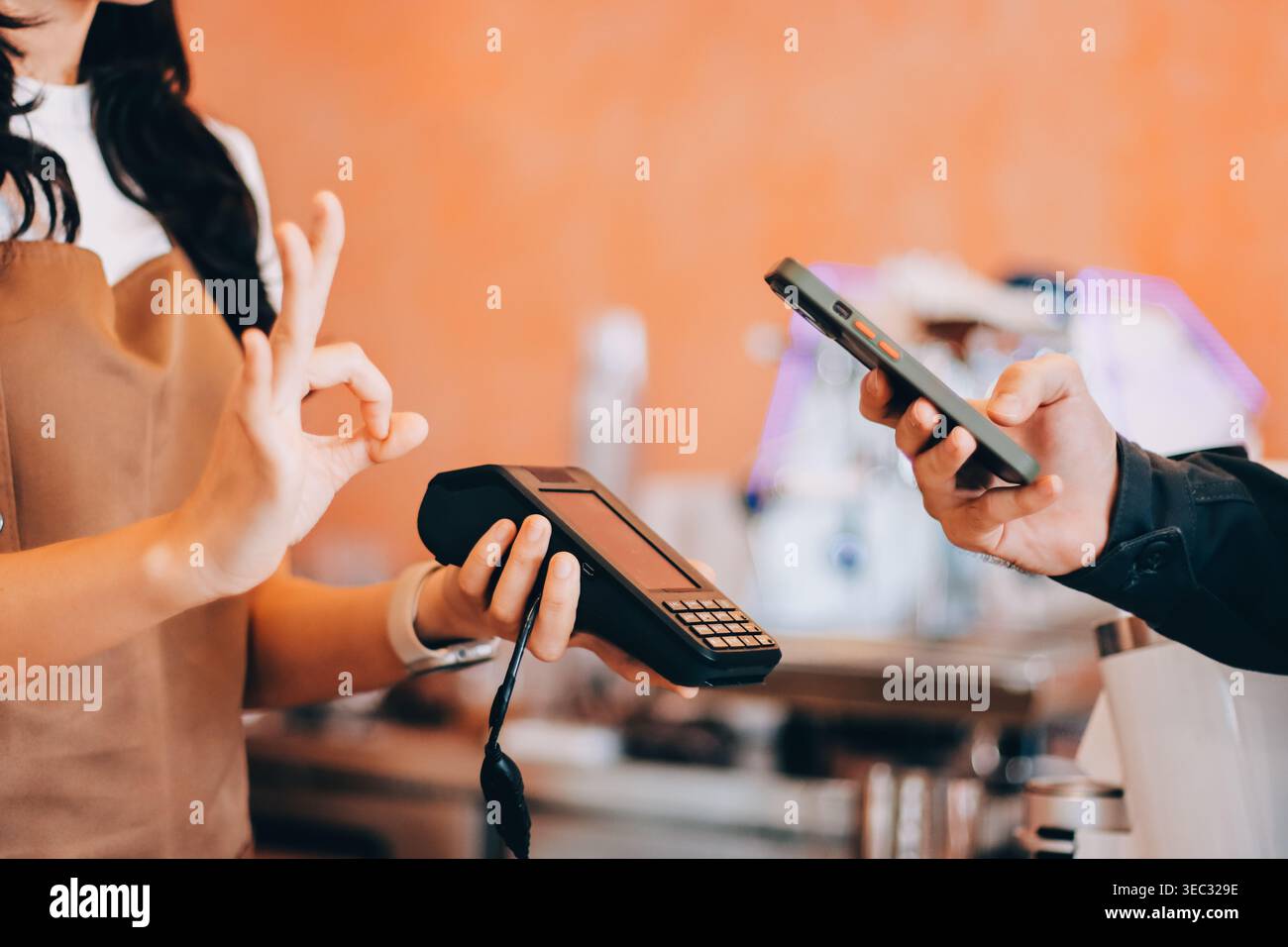 Barista asiatica anziana che riceve pagamenti mobili da un cliente tramite smartphone presso un bar che dimostra una moderna transazione senza contanti in un ambiente accogliente Foto Stock