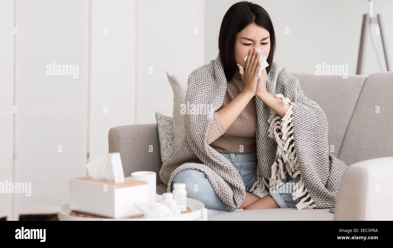 Donna seduta sul divano che si sente malata e starnuto mentre è avvolta in coperta Foto Stock