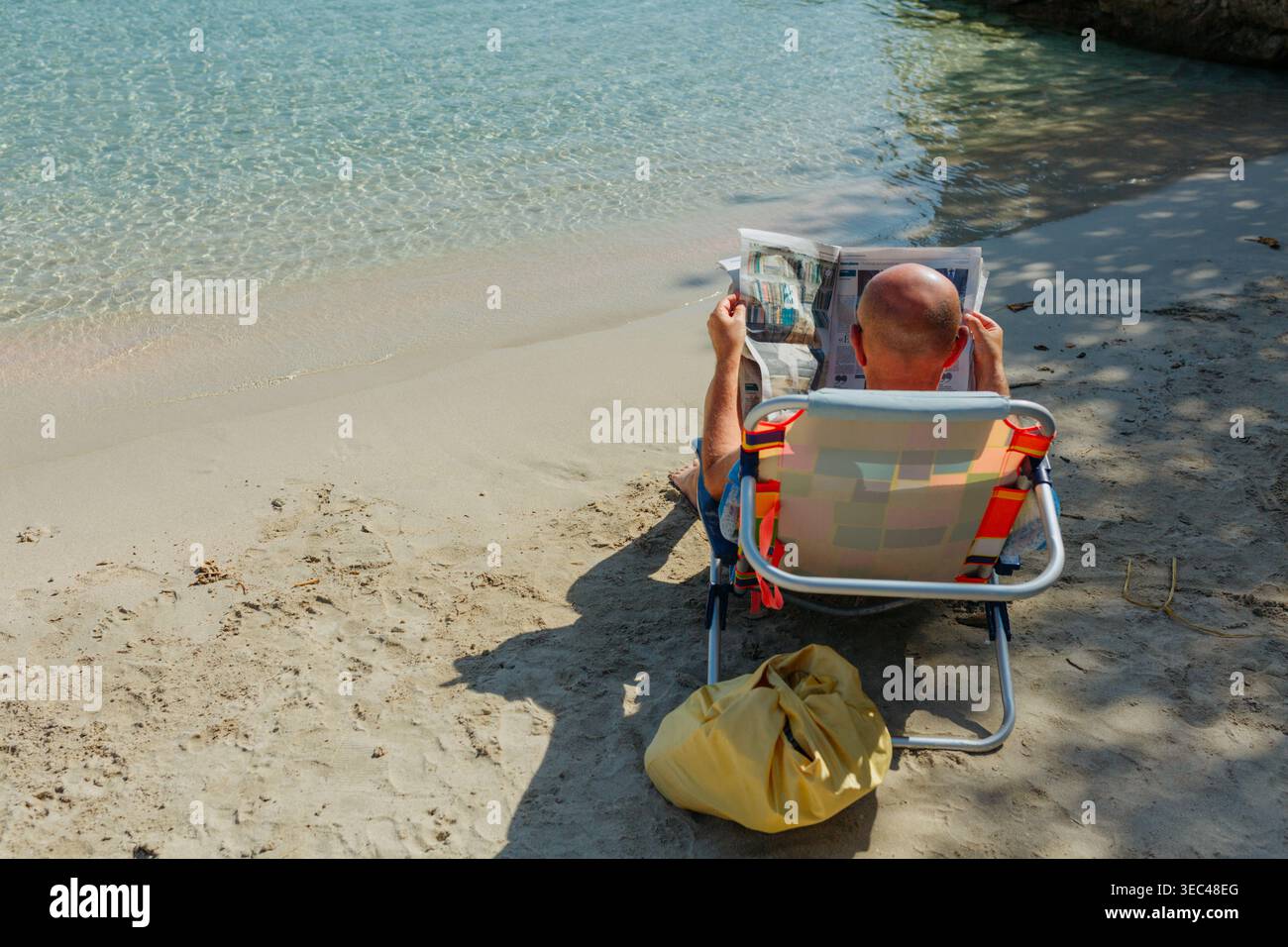 Vista posteriore di un uomo che legge il giornale sulla sedia da spiaggia sulla Sunny Tropical Shore Foto Stock