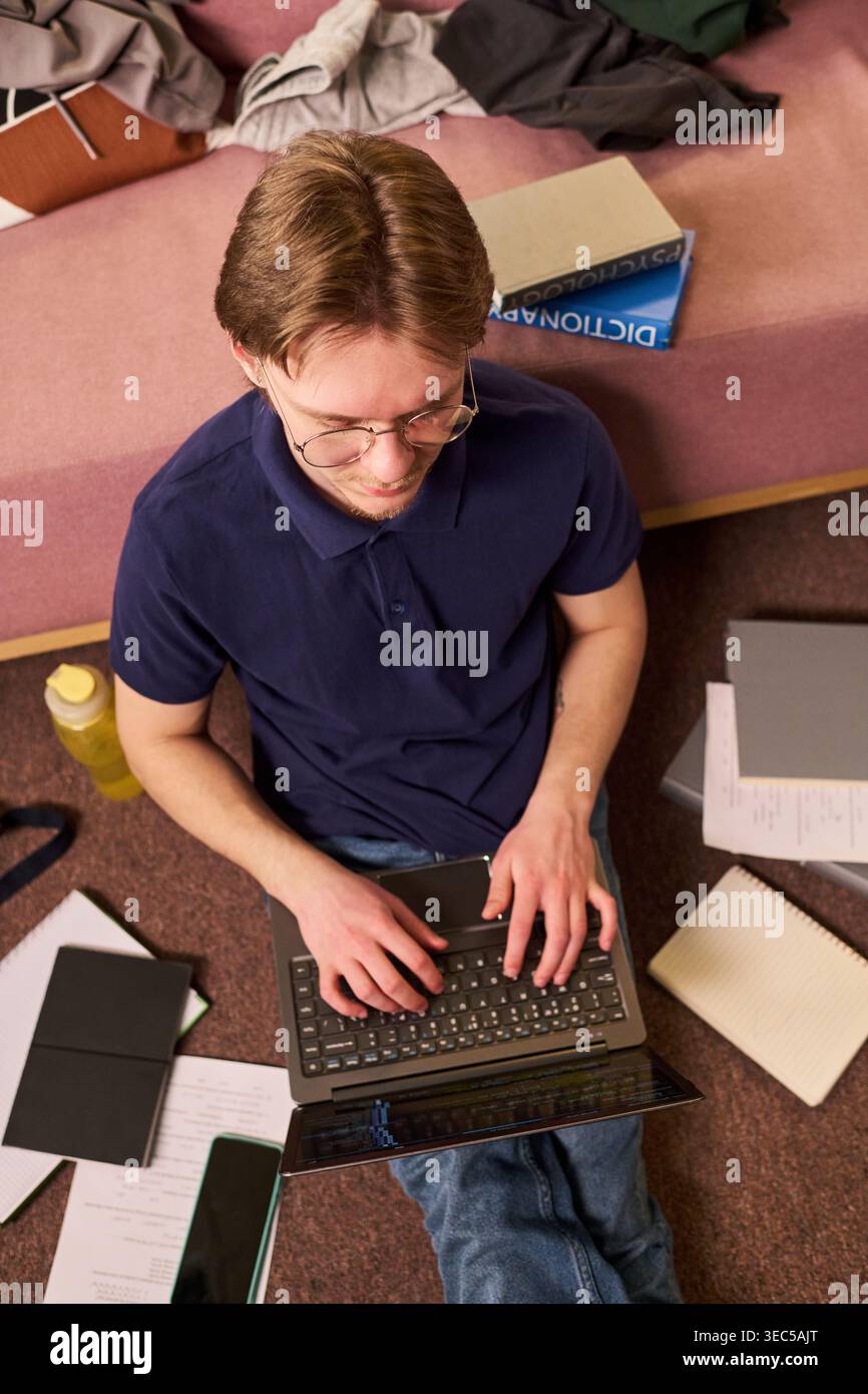 Ripresa verticale di uno studente caucasico maschio che scrive sul laptop mentre si siede sul pavimento accanto ai materiali di studio, preparando il corso. Utile per il marketing dell'istruzione, la vita universitaria, l'apprendimento online Foto Stock