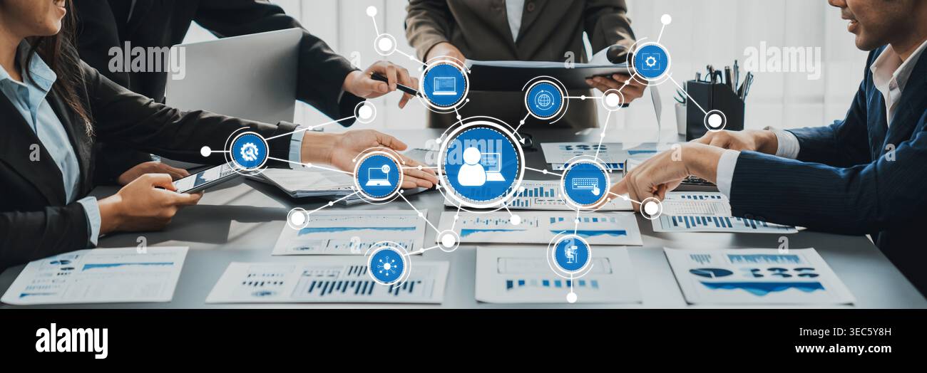 Un team di professionisti collabora attorno a un tavolo da riunione pieno di grafici e simboli digitali, sottolineando il lavoro di squadra e la pianificazione strategica per Foto Stock