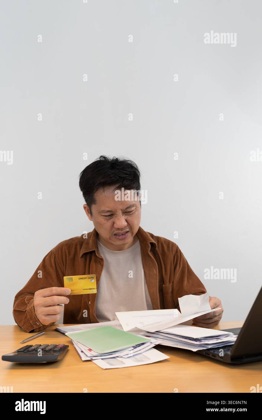 Un uomo preoccupato controlla i documenti mentre tiene una carta di credito alla sua scrivania, descrivendo debiti, pagamenti di bollette, pressioni sul budget, problemi finanziari personali e. Foto Stock
